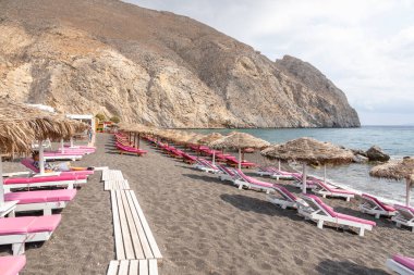Yunanistan 'ın Santorini adasındaki Perissa plajının üst manzarasında pembe güneşlikler ve şemsiyeler var. Plaj siyah, volkanik kumlarla kaplı, Yunanistan.