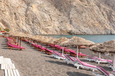 Yunanistan 'ın Santorini adasındaki Perissa plajının üst manzarasında pembe güneşlikler ve şemsiyeler var. Plaj siyah, volkanik kumlarla kaplı, Yunanistan.