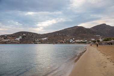 Paralia Gialos, Ios Adası, Yunanistan - 19 Eylül 2020: Kumlu sahil manzarası ve tepelerdeki geleneksel beyaz binalar. Körfezin üzerinde gün batımı.