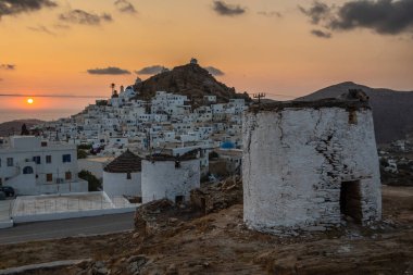 Chora 'nın ana meydanında ikonik, geleneksel yel değirmenleri. Sarı günbatımı. Tepenin arkasında küçük şapeller var. Chora, Ios Adası, Yunanistan.