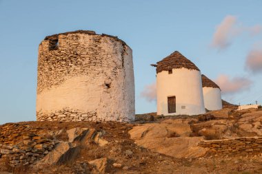 Chora 'nın ana meydanında ikonik, geleneksel yel değirmenleri. Sarı günbatımı. Tepenin arkasında küçük şapeller var. Chora, Ios Adası, Yunanistan.