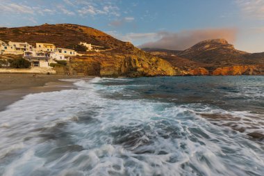 Agkali Sahili, Folegandros Adası, Yunanistan - 24 Eylül 2020: kumlu bir plajın yanındaki beyaz yamaç villaları. Turistler denizde yüzüyor ve sahilde dinleniyorlar. Gün batımı.