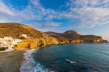Agkali Sahili, Folegandros Adası, Yunanistan - 24 Eylül 2020: kumlu bir plajın yanındaki beyaz yamaç villaları. Turistler denizde yüzüyor ve sahilde dinleniyorlar. Gün batımı.