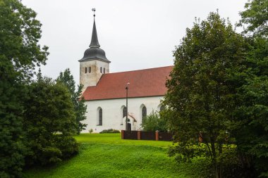 Estonya 'nın Viljandi şehrinde bulutlu bir yaz gününde yeşil ağaçlar ve çimlerle çevrili yüksek kulesi ve kırmızı çatısı olan beyaz ortaçağ kilisesi..