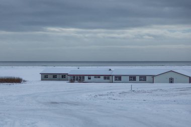 İzlanda 'da Krysuvik yakınlarında karla kaplı binalar sessiz bir kıyı şeridinde duruyor. Kış manzarası sakin deniz, bulutlu gökyüzü ve açık donmuş arazi.