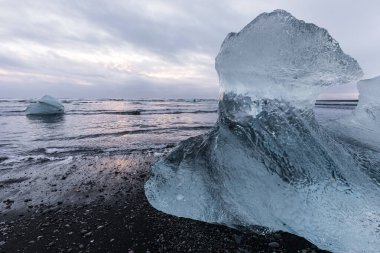 İzlanda 'daki Jokulsarlon Buzul Gölü' nün kara kum kıyısında dalgalar ve yüzen buzlarla kaplı büyük, yontulmuş bir buz kütlesi..