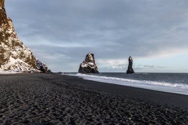 Reynisfjara siyah kumsal İzlanda 'da bazalt deniz yığınları, Atlantik dalgaları ve dramatik gökyüzü altında kış kıyı manzarası.