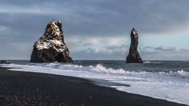 Reynisfjara siyah kumsal İzlanda 'da bazalt deniz yığınları, Atlantik dalgaları ve dramatik gökyüzü altında kış kıyı manzarası.