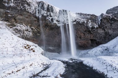 İzlanda 'daki Seljalandsfoss şelalesi kış boyunca karla kaplı uçurumlar ve buzla kaplı çevresiyle donmuş bir nehre akıyor..