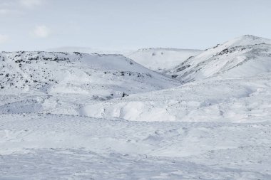 İzlanda, Reykjadalur Vadisi 'nde karla kaplı dağlar ve volkanik arazi. Donmuş akıntılı kış manzarası, engebeli tepeler ve bulutlu gökyüzü altında el değmemiş İskandinav doğası.