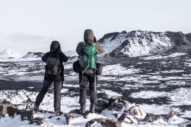 Grindavik, Reykjanes Yarımadası, İzlanda 6 Şubat 2022: Karda duran iki yürüyüşçü kayaları kapladı ve kış koşullarında dağlarla kaplı engebeli bir volkanik lav alanını gözlemledi.