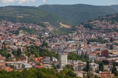 Saraybosna, Bosna-Hersek: 15 Ağustos 2022: Çağdaş ofis kuleleri, yerleşim bölgeleri ve arka planda yeşil tepeler bulunan Saraybosna şehir merkezinin panoramik hava manzarası.