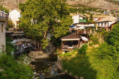 Bosna-Hersek 'in Mostar kenti 16 Ağustos 2022: Eski Mostar kentinin çatılarından yükselen bir cami minaresi olan geleneksel taş binadaki restoran teraslarında oturan insanlar.