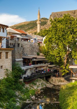 Bosna-Hersek 'in Mostar kenti 16 Ağustos 2022: Eski Mostar kentinin çatılarından yükselen bir cami minaresi olan geleneksel taş binadaki restoran teraslarında oturan insanlar.