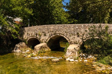 Bosna-Hersek 'in Stolac kentinde, güneşli bir yaz gününde ağaçlar ve yeşilliklerle çevrili sığ bir nehri kaplayan eski taş köprü..