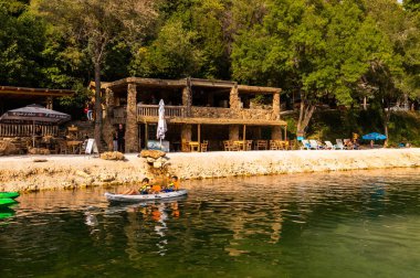 Kravica Şelalesi, Bosna ve Hercegovina - 18 Ağustos 2022, çocuklar yemyeşil ağaçları olan nehir kenarındaki taştan bir kafe yakınındaki sakin nehirde kayak yapıyorlar..