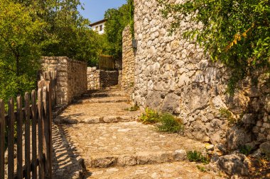 Geleneksel taş duvarlar ve Bosna-Hersek 'in güneş ışığı altındaki yemyeşil yeşilliklerle çerçevelenmiş tarihi bir köy caddesine çıkan yokuş yukarı merdiven..