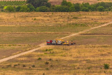 Ravno, Bosna ve Hercegovina - 19 Ağustos 2022: Kuru bir alana park edilmiş inşaat makineleri, bulutlu bir gökyüzünün altında toprak bir yolda bekliyor.