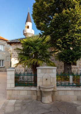 Bosna-Hersek 'in Trebinje kentinde geleneksel bir halk çeşmesinin yer aldığı taş duvarların üzerinden yükselen Osman Paşa camii minaresi.