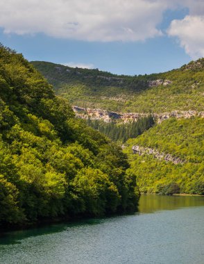 Trebisnjica nehir suyu, Bosna-Hersek 'in Trebinje kentinde parlak bir yaz gökyüzü altında yemyeşil orman tepeleri ve kayalık kayalıkları yansıtıyor.