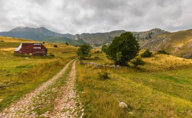 Bosna-Hersek 'in Lukomir kentindeki bulutlu gökyüzünün altında dolambaçlı bir yol, eski bir geleneksel kulübe ve yuvarlanan tepelere ev sahipliği yapan kırsal dağ manzarası.
