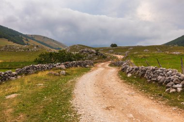 Bosna-Hersek 'in bulutlu gökyüzünün altındaki engin yeşil bir dağ manzarasında geleneksel kuru taş duvarlarla çevrili, dolambaçlı toprak yol.