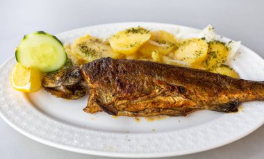 Kızarmış alabalık, pişmiş patates, taze soğan dilimleri, limon dilimi ve beyaz tabakta salatalık ile servis edilir..