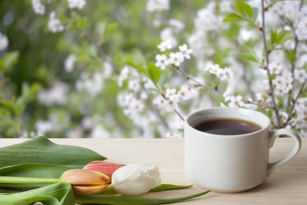 Kawa w filiżance i tulipany na drewnianym stole naprzeciwko rozmyte — Zdjęcie stockowe