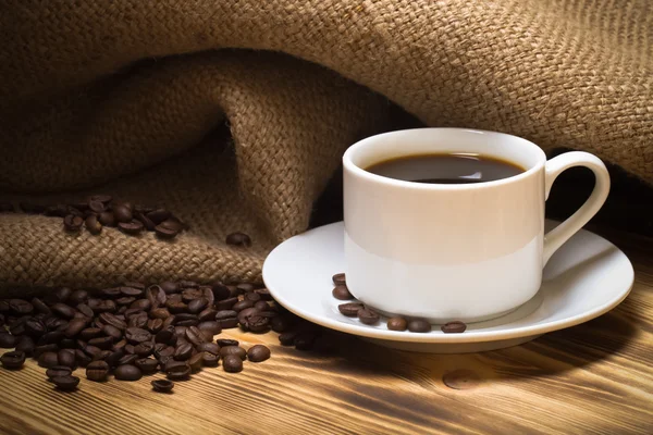 Grãos de café e café em copo branco na mesa de madeira com serapilheira — Fotografia de Stock