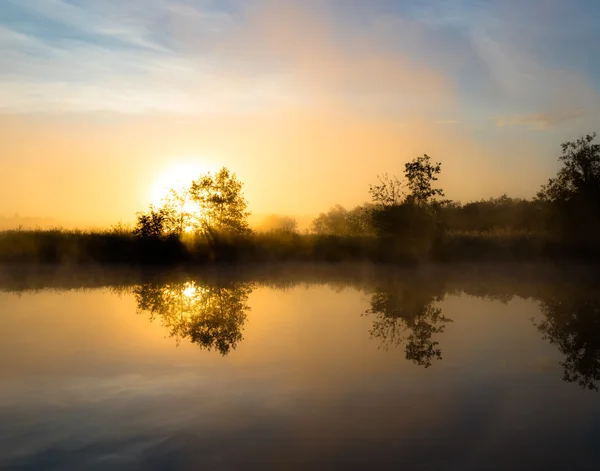 Mlžné svítání nad řekou. Sluneční paprsky zpoza stromu — Stock fotografie