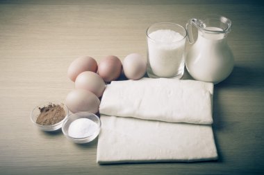 Süt, şeker, kakao, puf hamur, yumurta ve tuz bir ahşap tahta üzerinde.