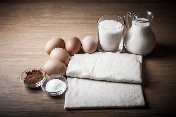 Mléko, cukr, kakao, listového těsta, vejce a sůl na dřevěném prkénku. — Stock fotografie