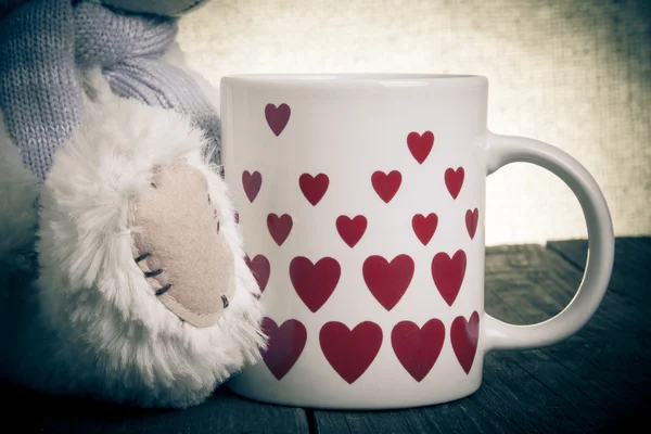 Paw of Teddy bear  and mug with hearts on the old wooden table. — Stock Photo, Image