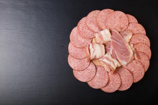 Slices of sausage and bacon on a round cutting board on a black — Stock Photo, Image