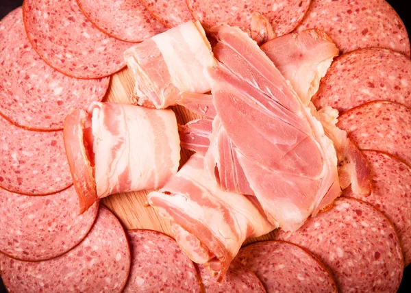 Slices of sausage and bacon on a round cutting board. Toned — Stock Photo, Image