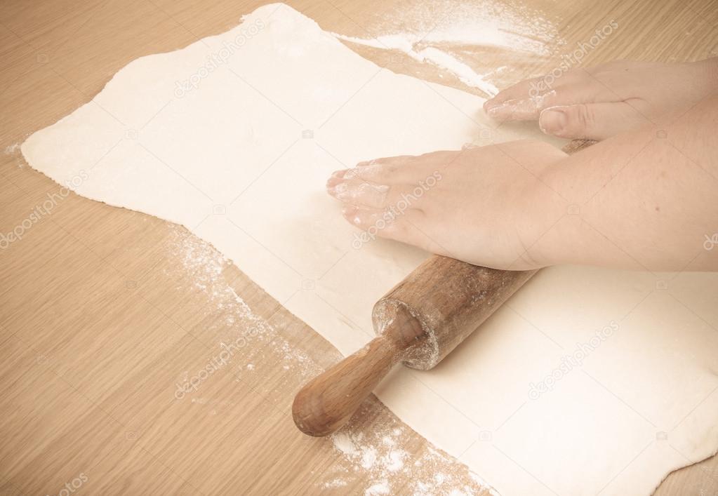 Plump women's hands roll out the dough on a light wooden table. 