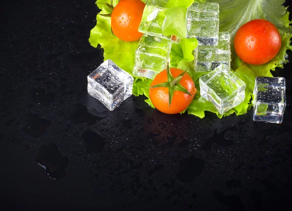 Röda körsbär tomater, grön sallad och isbitar på svart våt bord — Stockfoto