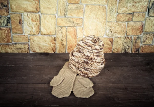 Knited hat and mittens on wooden table opposite a stony wall. To — 图库照片