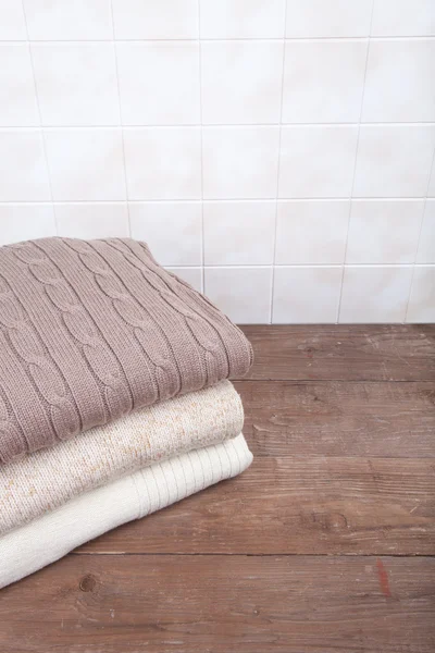 Stack of knitted clothes on wooden table opposite a tile wall — ストック写真