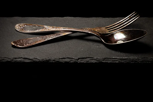 Tenedor y cuchara en un plato de pizarra sobre un fondo negro. Tonificado —  Fotos de Stock