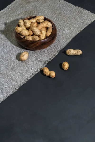 Leguminous plant is a source of vitamins, antioxidants, iron, hemoglobin and fiber, nut contains niacin B3, peanuts in shell in a wooden bowl on a table on a black concrete background with copy space