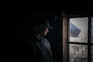 Karanlık ve korku dolu, terk edilmiş bir evin karanlık tavan arasında pencereden dışarı bakan şüpheli bir adamın silüeti.