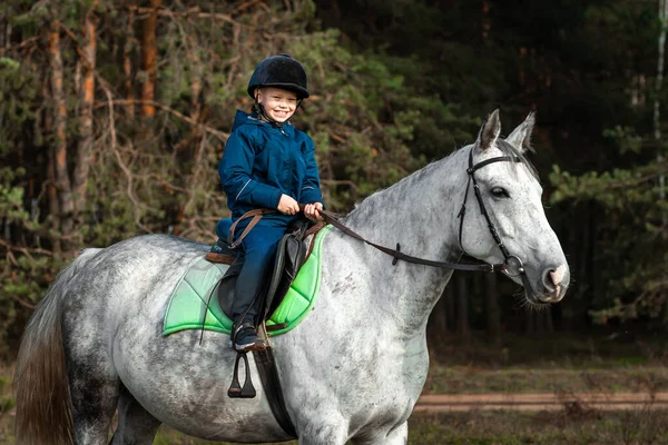 Little boy in a jockey cap on a white adult horse on a background of nature. Jockey, hippodrome, horseback riding.