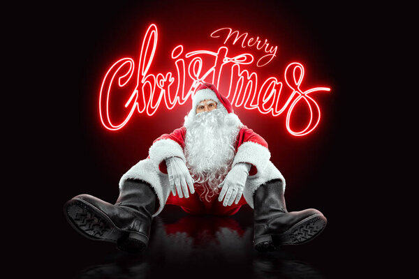 Portrait of Santa Claus sitting in a red suit against the backdrop of neon lettering merry christmas, black background. Concept for Christmas Eve, Vacation, Holiday Banner, New Year