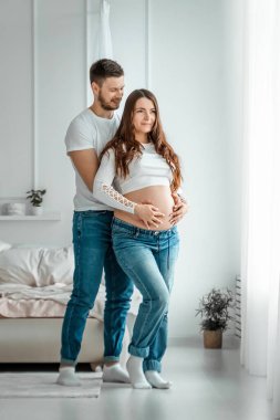 Kocasıyla birlikte hamile bir kız parlak bir yatak odasında duruyor. Genç, çekici, hamile bir kızın güzel göbeği. Aile, evlilik, doğum kavramı