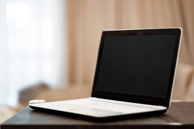 Bulanık bir odanın arka planına karşı masada açısal pozisyonda siyah ekranlı bir dizüstü bilgisayar. İş yeri, ofis, birlikte çalışma kavramı.