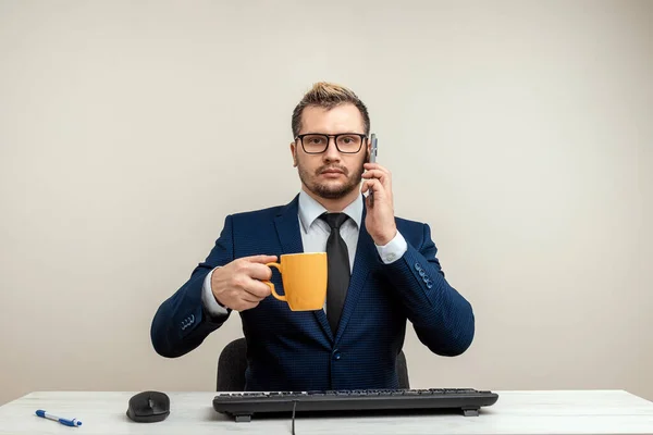 A businessman in a blue suit at a work table. Holds a mug and a phone. Emotional work. Multitasking, effective business worker concept