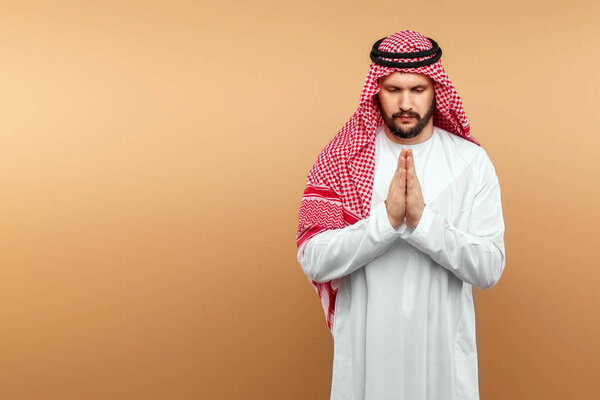 Arab man businessman in national clothes folded his hands in prayer, like, beige background. Dishdasha, kandora, thobe, middle east traditional menswear concept, islam. Copy space