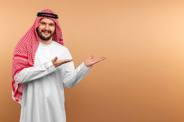 Arab male businessman in national clothes points with his hands to something, Beige background. Dishdasha, kandora, thobe, middle east traditional menswear concept, islam. Copy space
