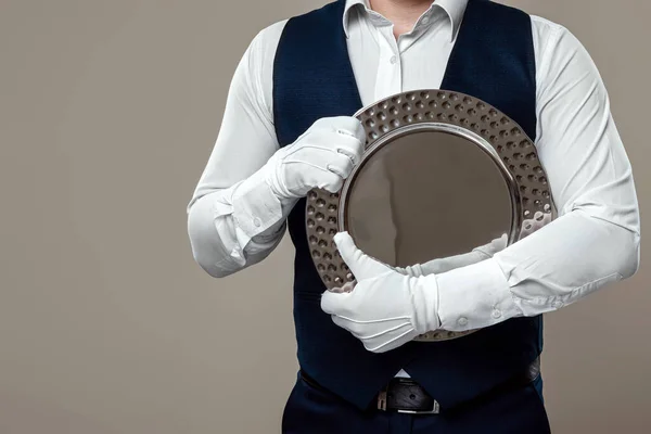 A male waiter in a white shirt and white gloves, standing with a silver tray, hugs it to his chest. The concept of serving staff, serving customers in a restaurant